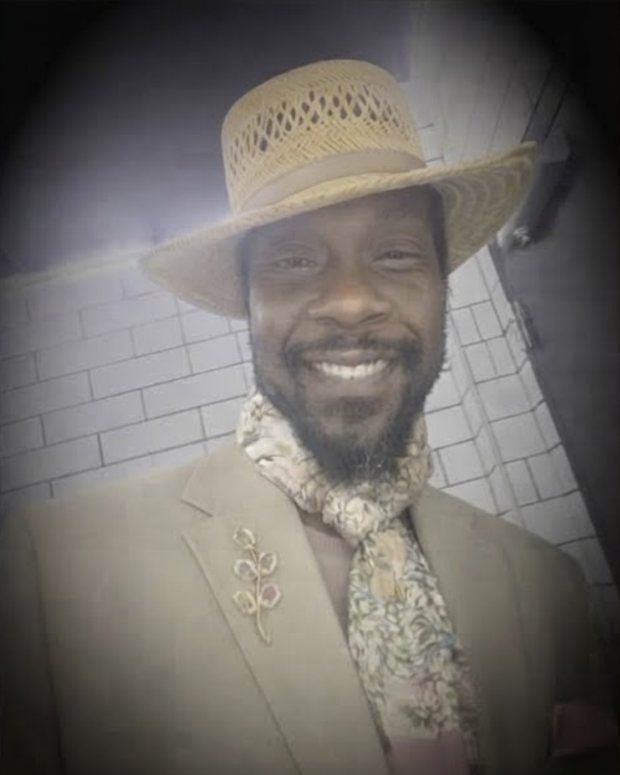 George Gentleman in a cream suit, straw boater, floral cravat and a gold lapel brooch